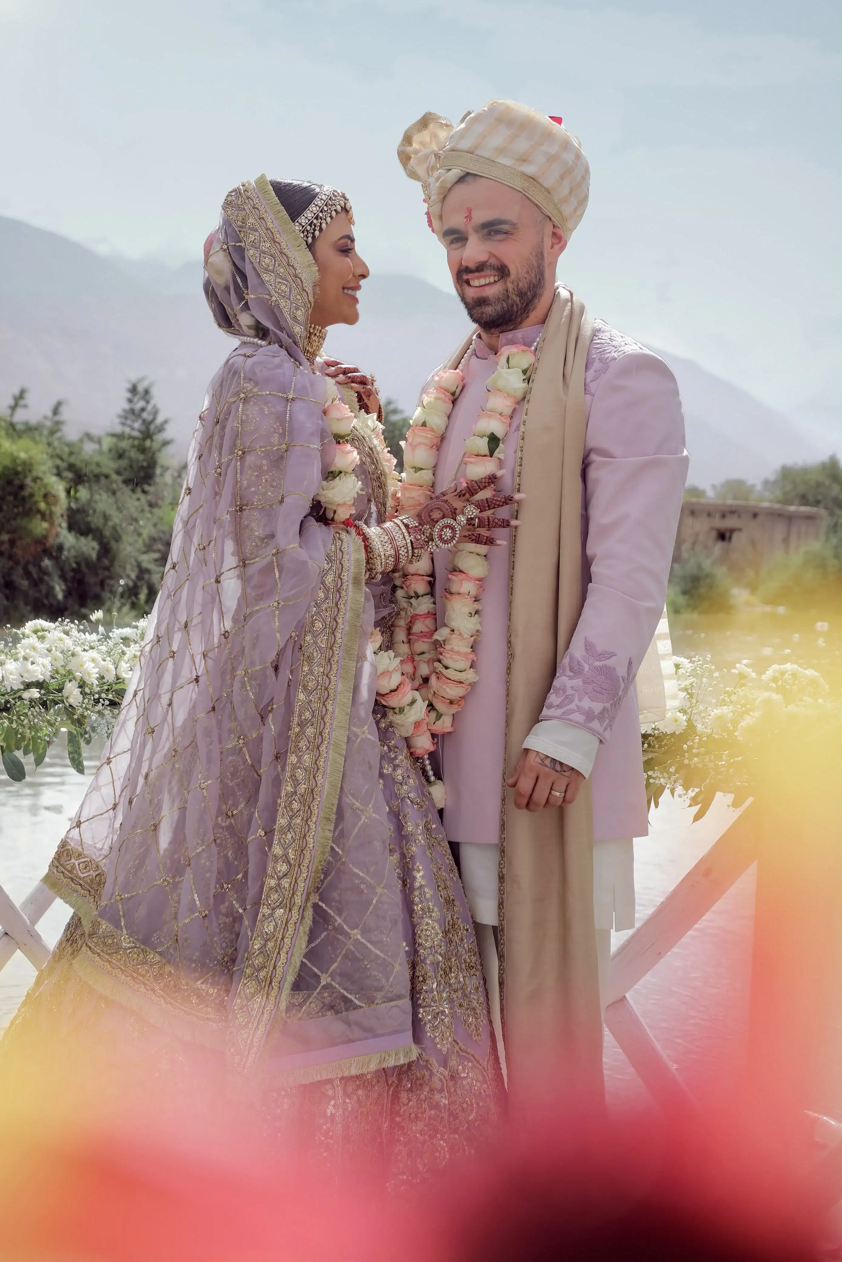 Bride and groom smiling with rose garlands by the water